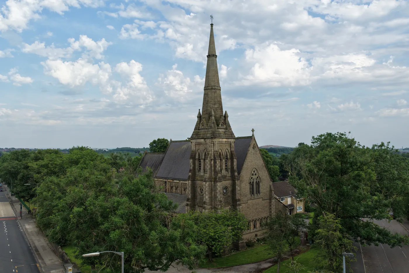 The spire of St Thomas rising from a canopy of summer trees beneath a wide, clouded sky