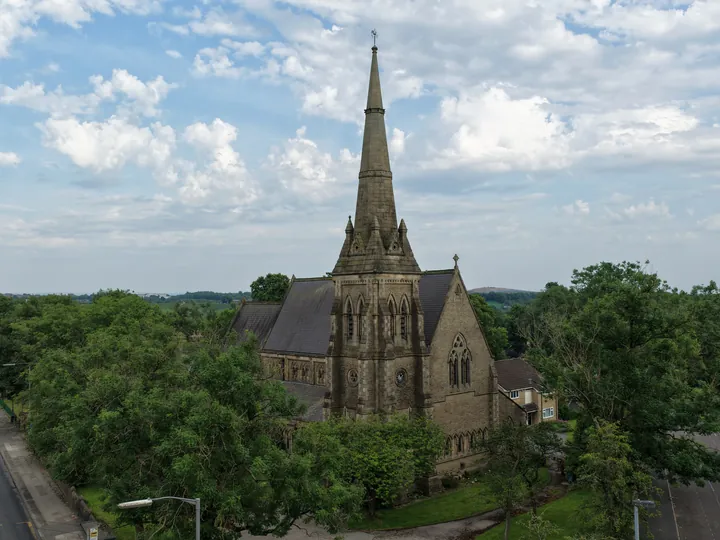 The spire of St Thomas rising from a canopy of summer trees beneath a wide, clouded sky